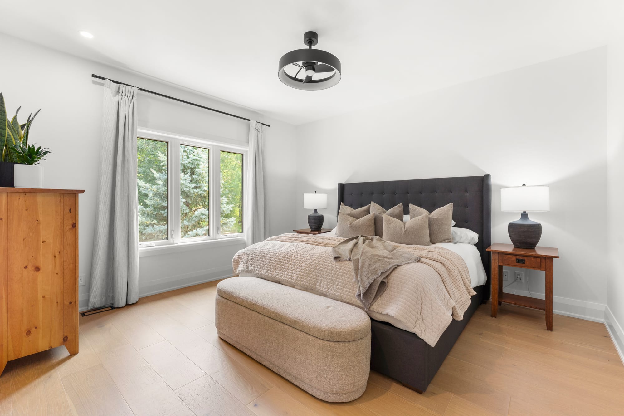 Primary bedroom with a tufted headboard, upholstered storage bench and a window looking onto mature evergreens