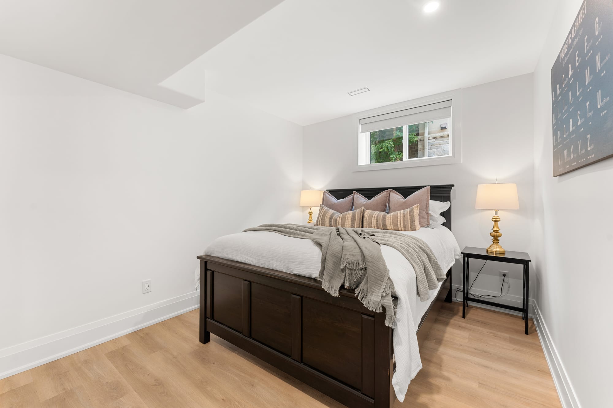 Second lower-level bedroom with a dark wood bed frame, brass lamps and a bright egress window